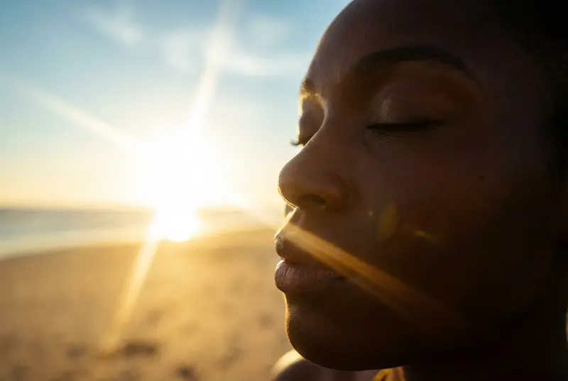 Close-up profile of a Black woman soaking up the sun with a bright lens flare in the background.