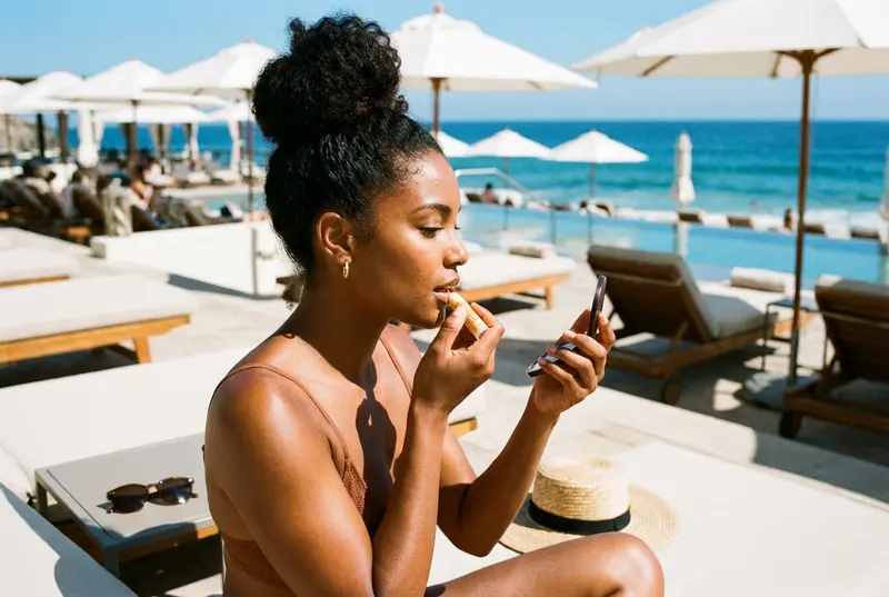 Woman applying protective lip balm while sitting on a beach lounge chair.
