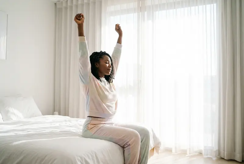 A woman waking up in a sunlit bedroom stretching and looking peaceful.